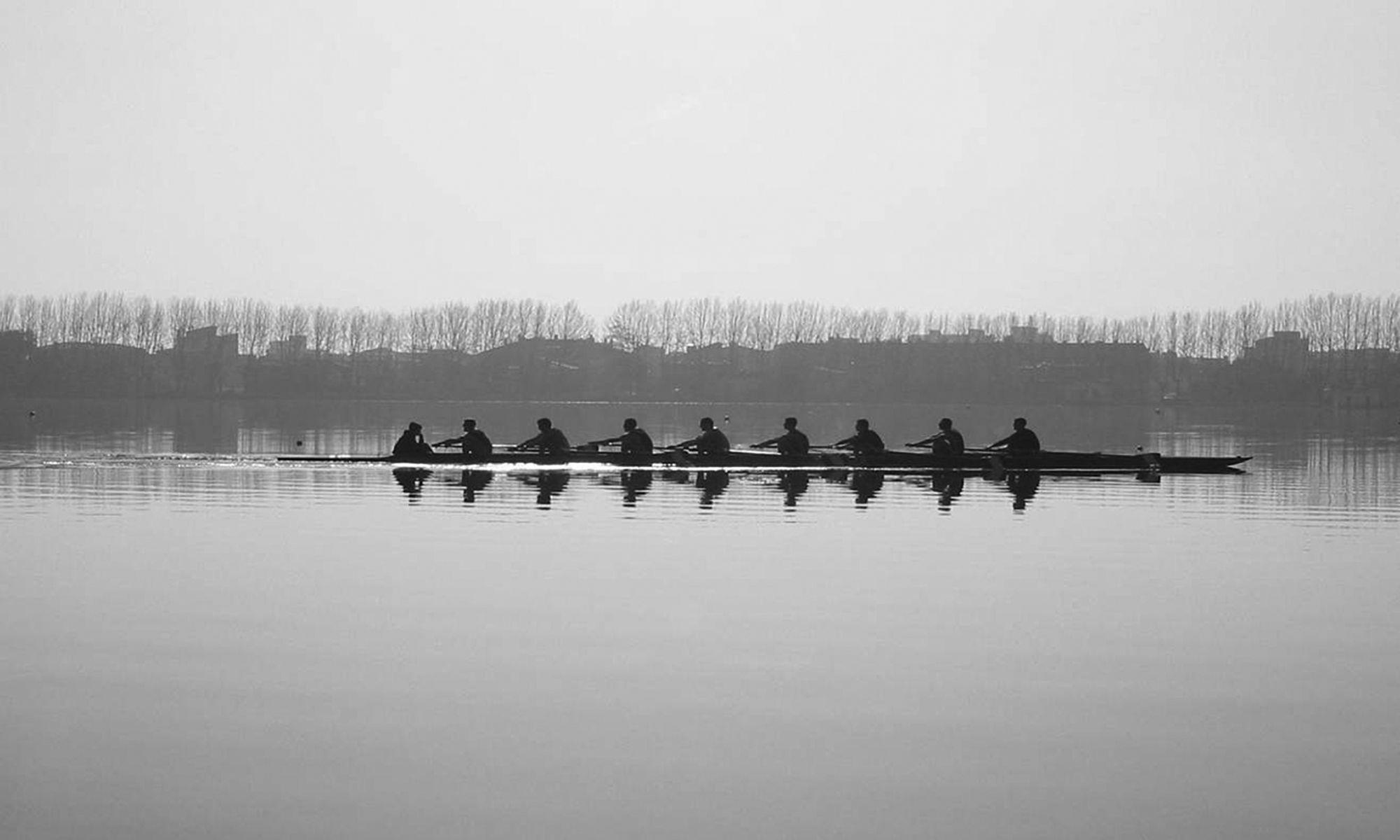 A black and white photo of nine people rowing a crew boat on a still lake.