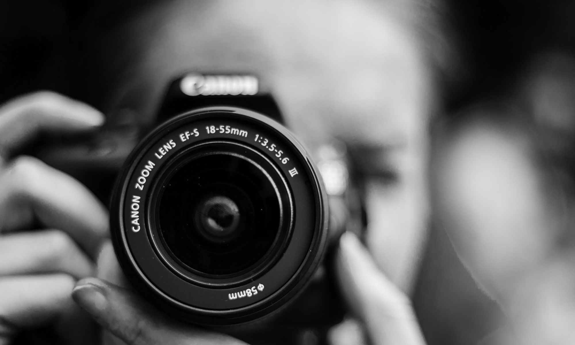 A black and white photo of a person pointing a camera directly ahead close up, with the camera lense in focus.